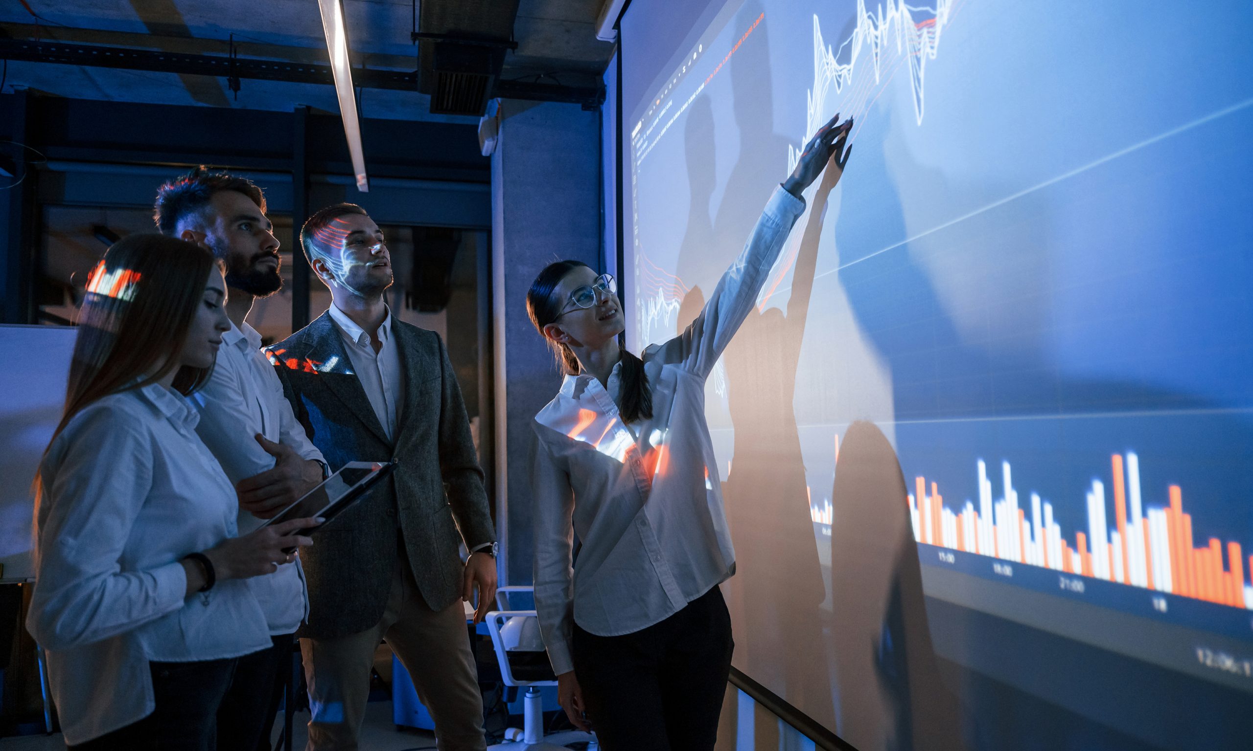 holding tablet. male leader talking to employees, showing the plan on the projector in office of stock exchange company
